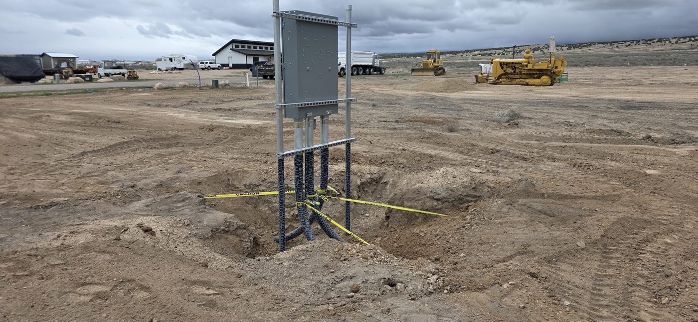 Large electrical service enclosure on a strut rack with multiple conduits, shop building and heavy equipment behind