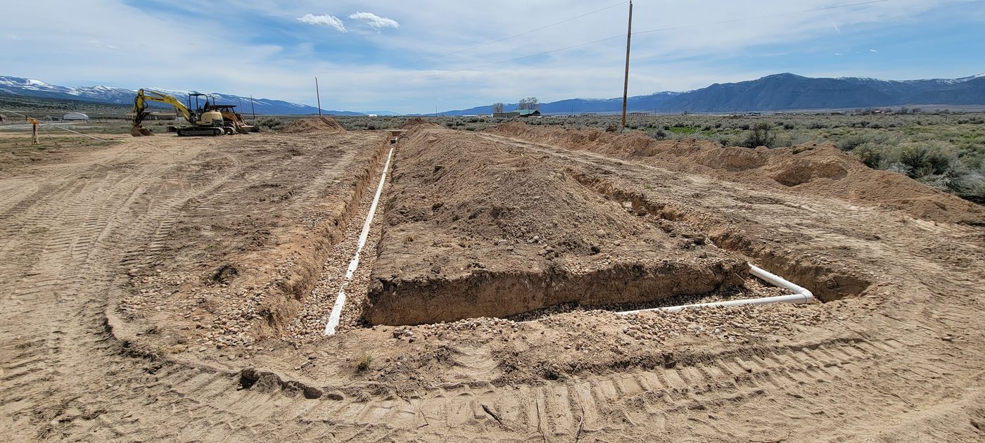 Drain field trenches cut into a sagebrush valley floor with an excavator and snow-capped mountains behind, rural Utah septic installation