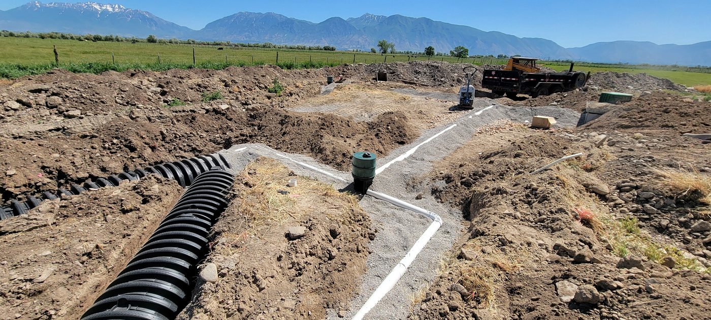 Rows of chamber drain field trenches with gravel and a distribution riser, mountains and pasture behind, Utah