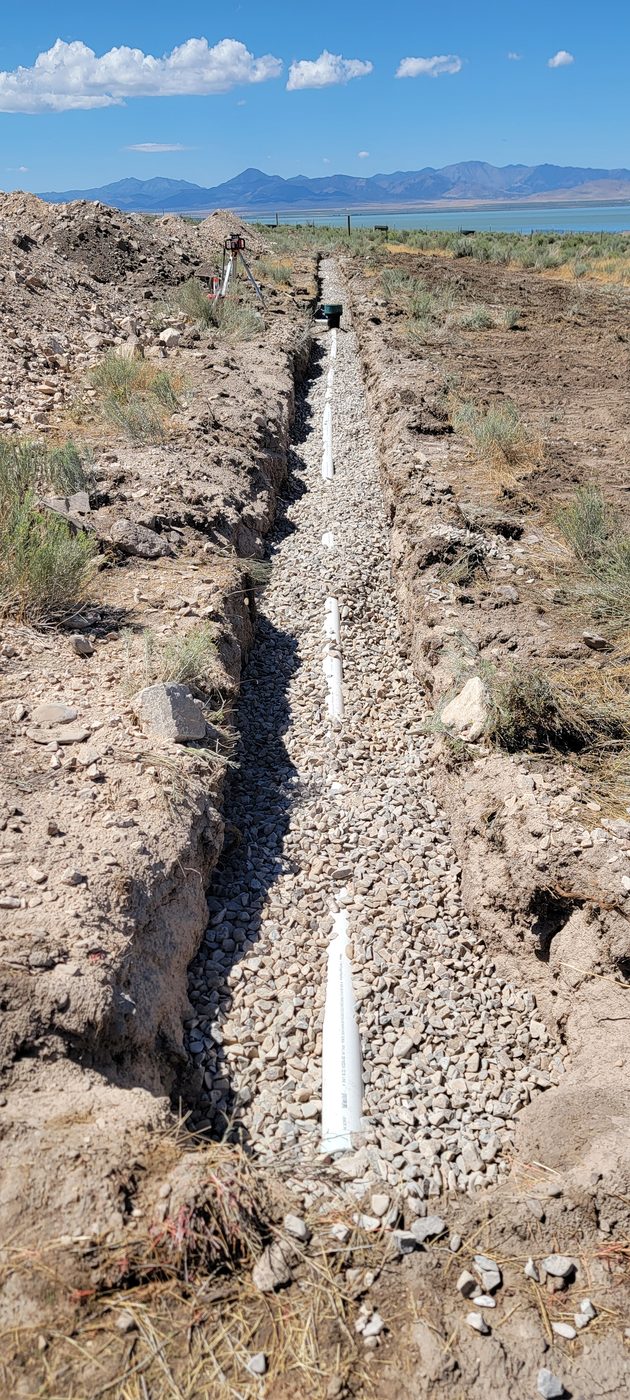 Long gravel-bedded drain field trench with perforated pipe and a laser level on a tripod, sagebrush and mountains beyond