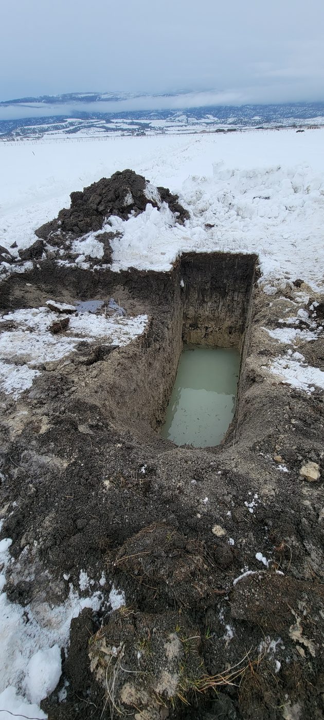 Soil evaluation pit where the water table was encountered, standing water in the bottom, snow-covered Utah valley behind