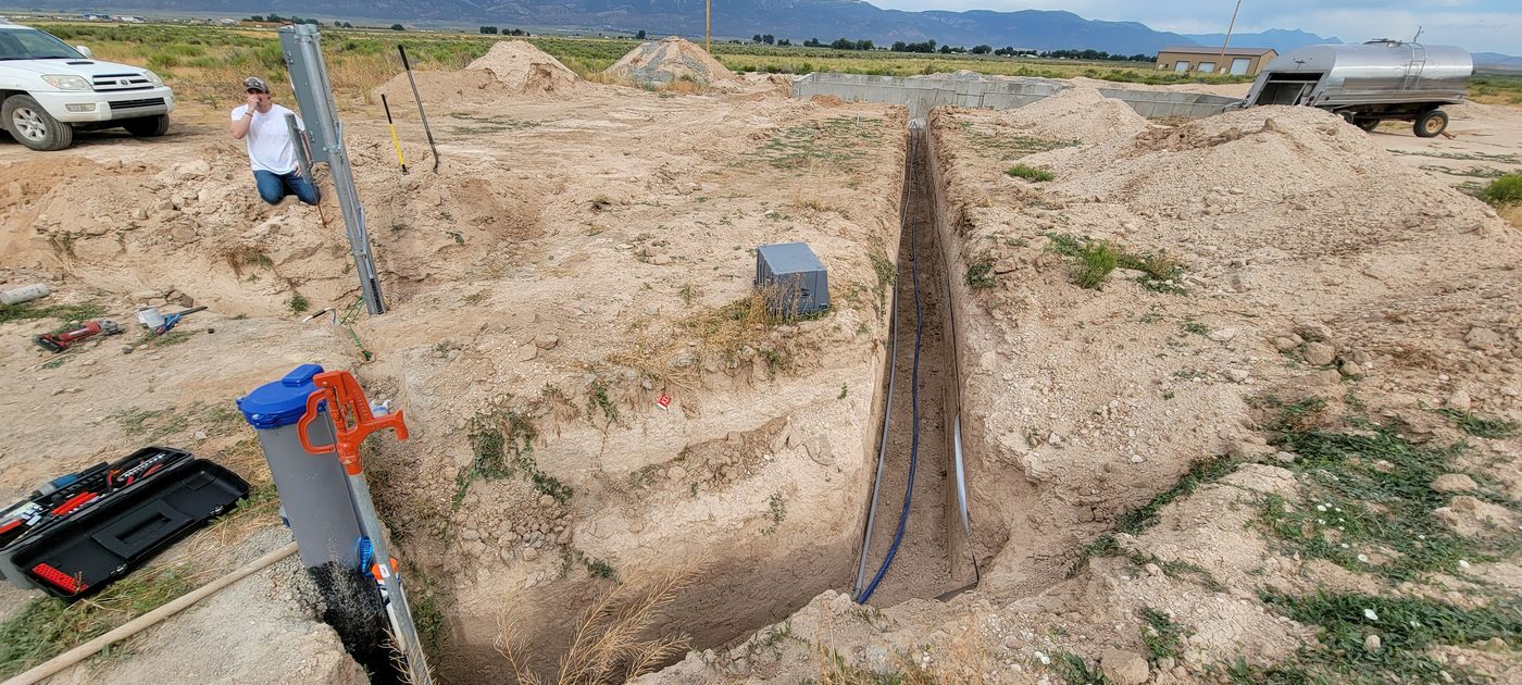 Long open trench with water service line and conduit running toward a foundation on a rural Utah property